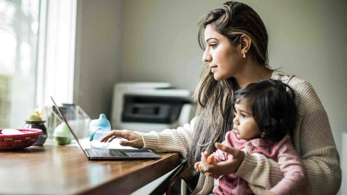 A woman sits at a table looking at a laptop with a child on her knee.