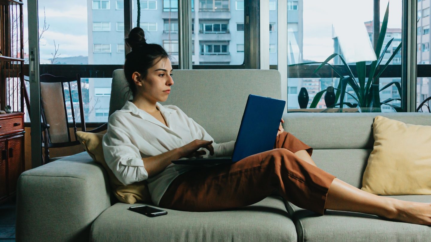 brunette woman in orange pants and a white blouse is sitting on her grey couch on her laptop, applying for a personal loan online during lockdown