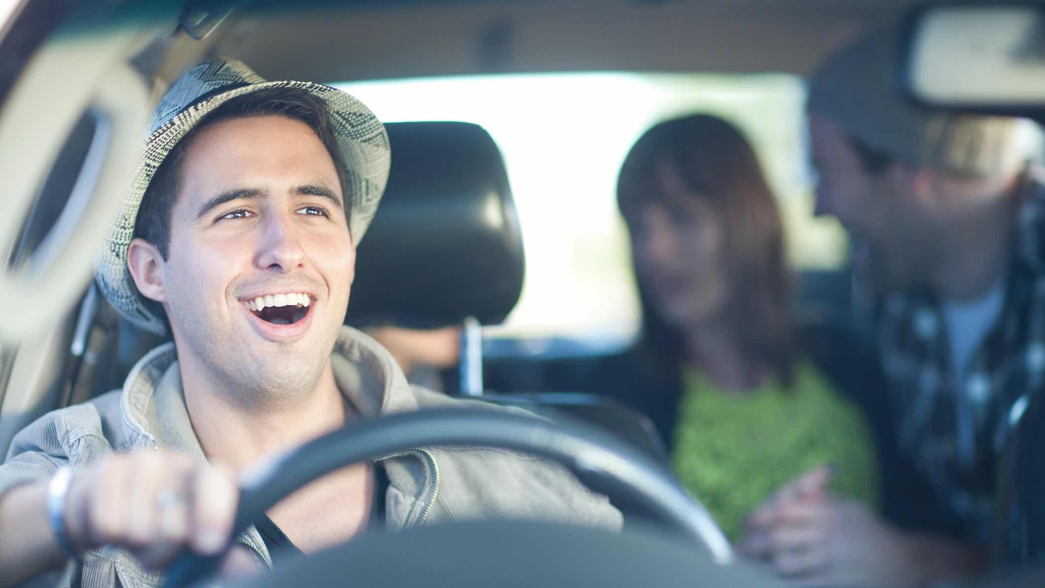 Rideshare driver in bucket hat smiling as passengers chat in the backseat.