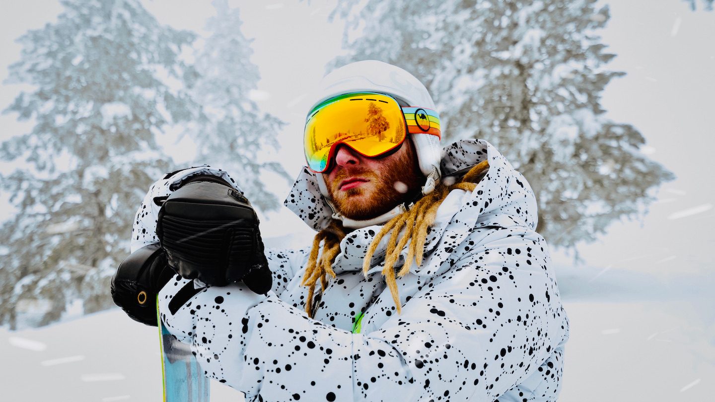 A ginger snowboarder in goggles, a white coat in the snowy, forested mountains