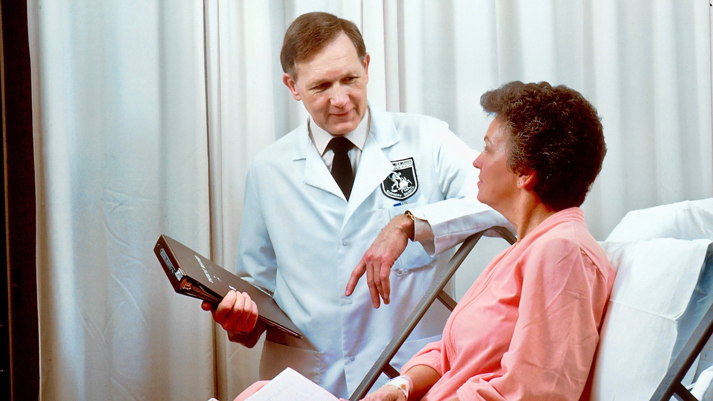 White male doctor consults a female patient dressed in pink while she sits upright in her hospital bed