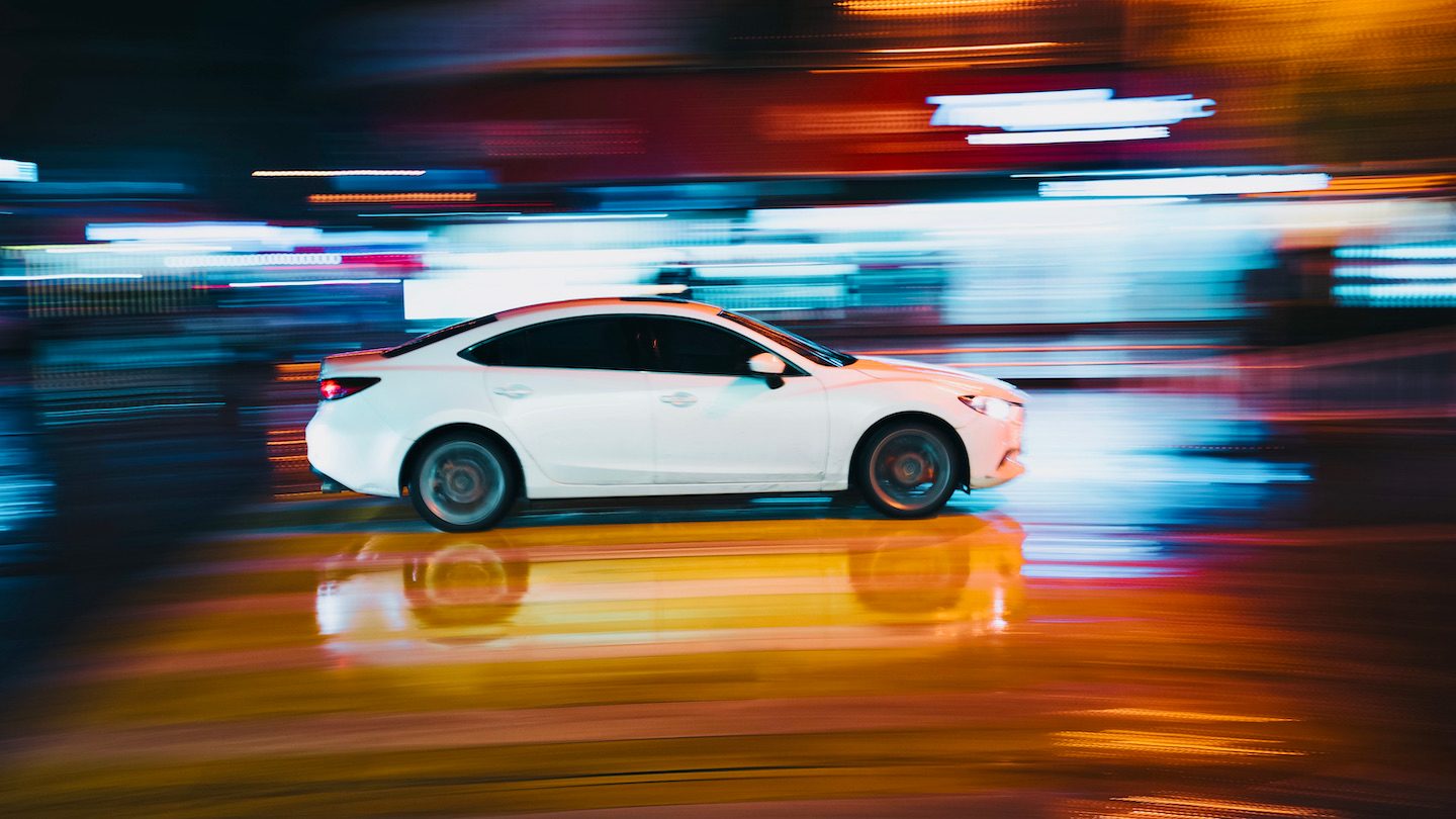 A white sedan speeds through a colourful motion blur street.