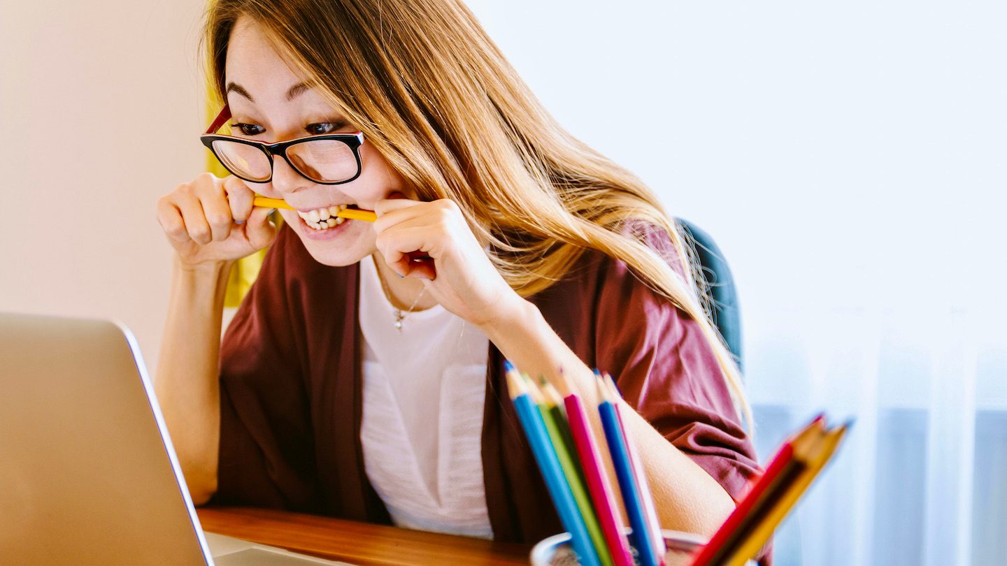 A woman bites down on a pencil in frustration while reading her laptop.