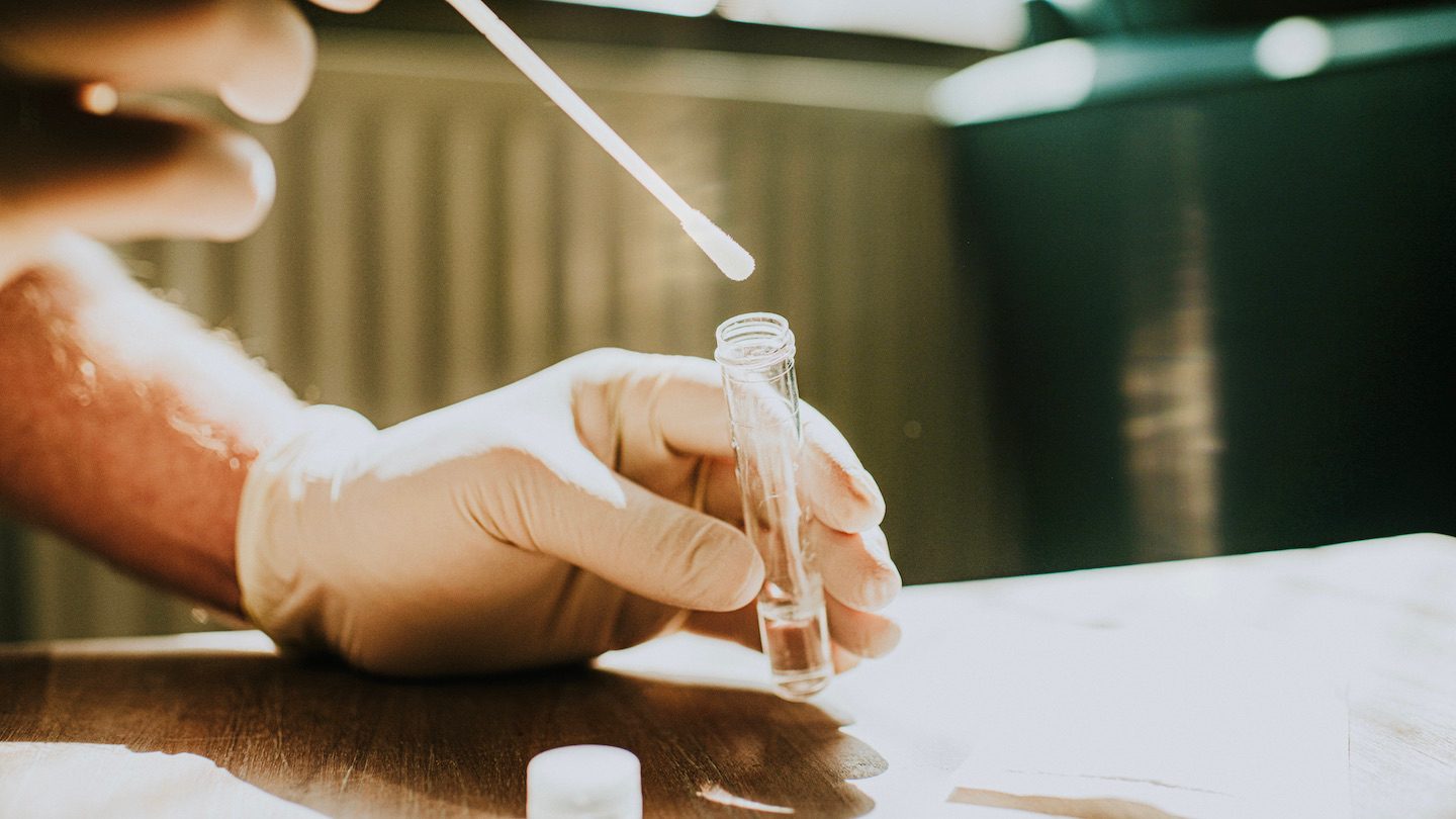 A latex gloved hand inserts a q tip into a COVID test tube.
