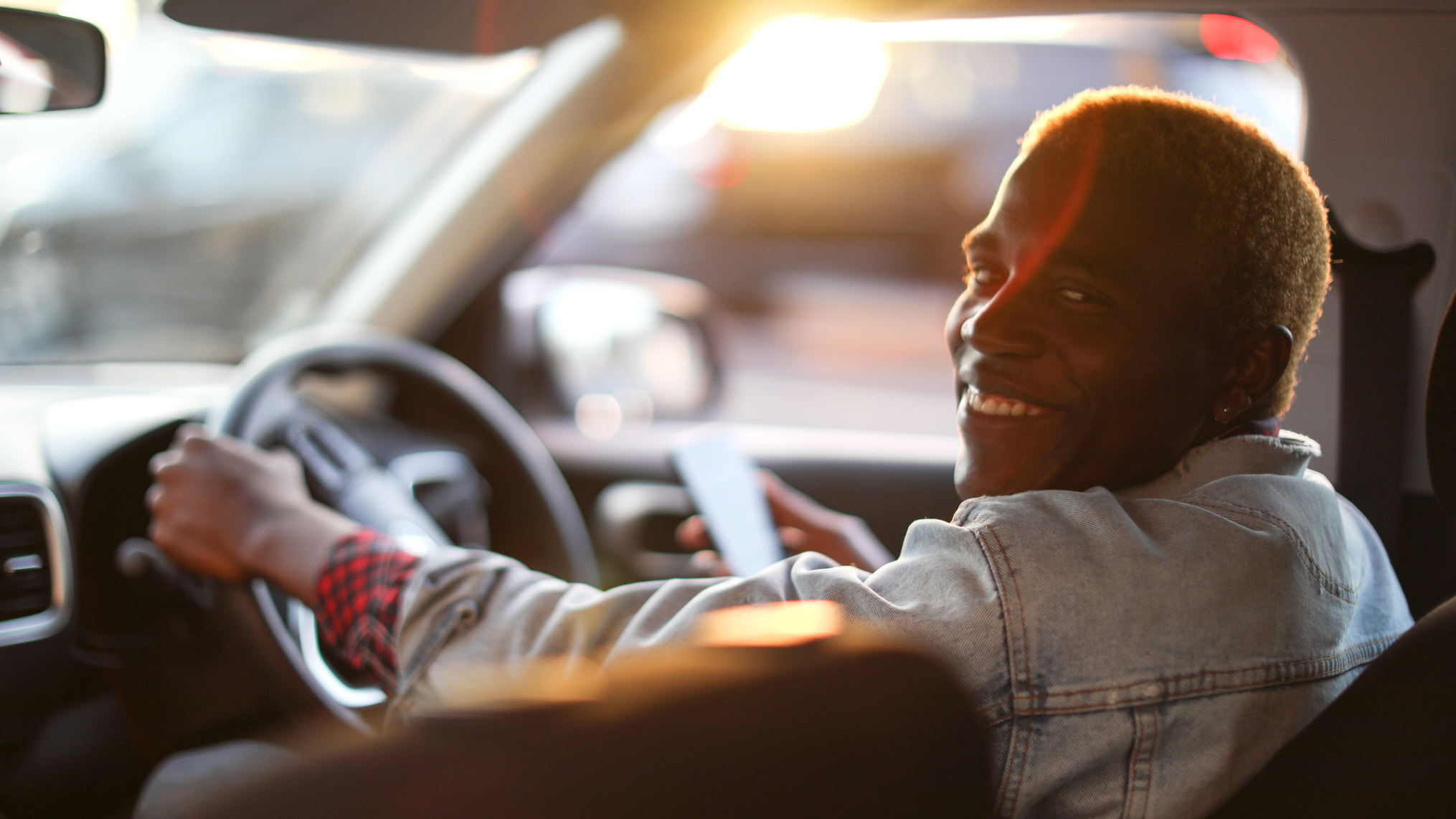 A man smiling at his back seat passengers from the driver's seat of his car. The sun sets in the distance.