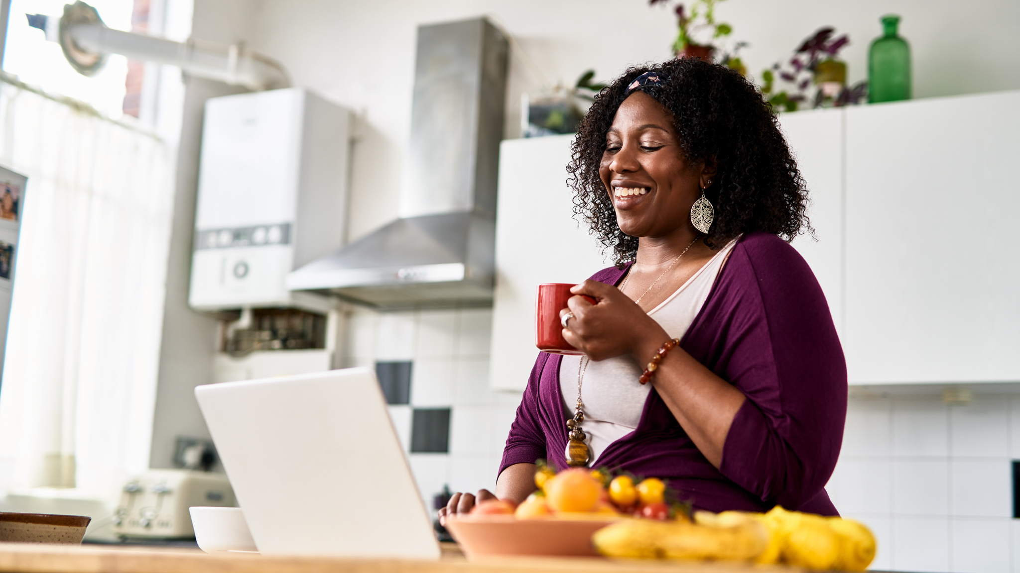 A woman, standing in her kitchen, smiles at her laptop. She is satisfied with her online shopping experience.