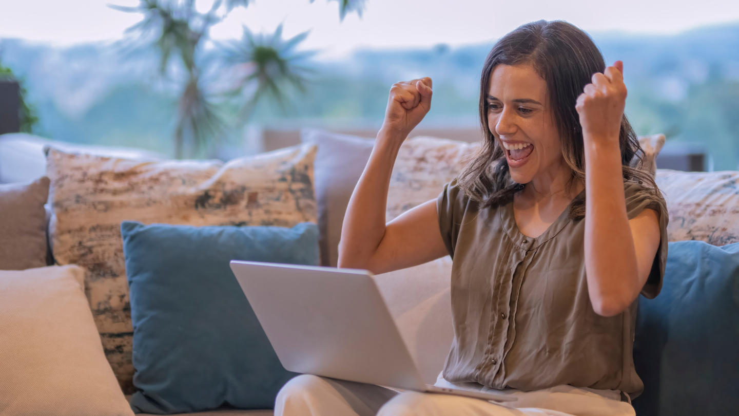 Young woman sitting on a sofa with a laptop celebrating getting a quiz question correct