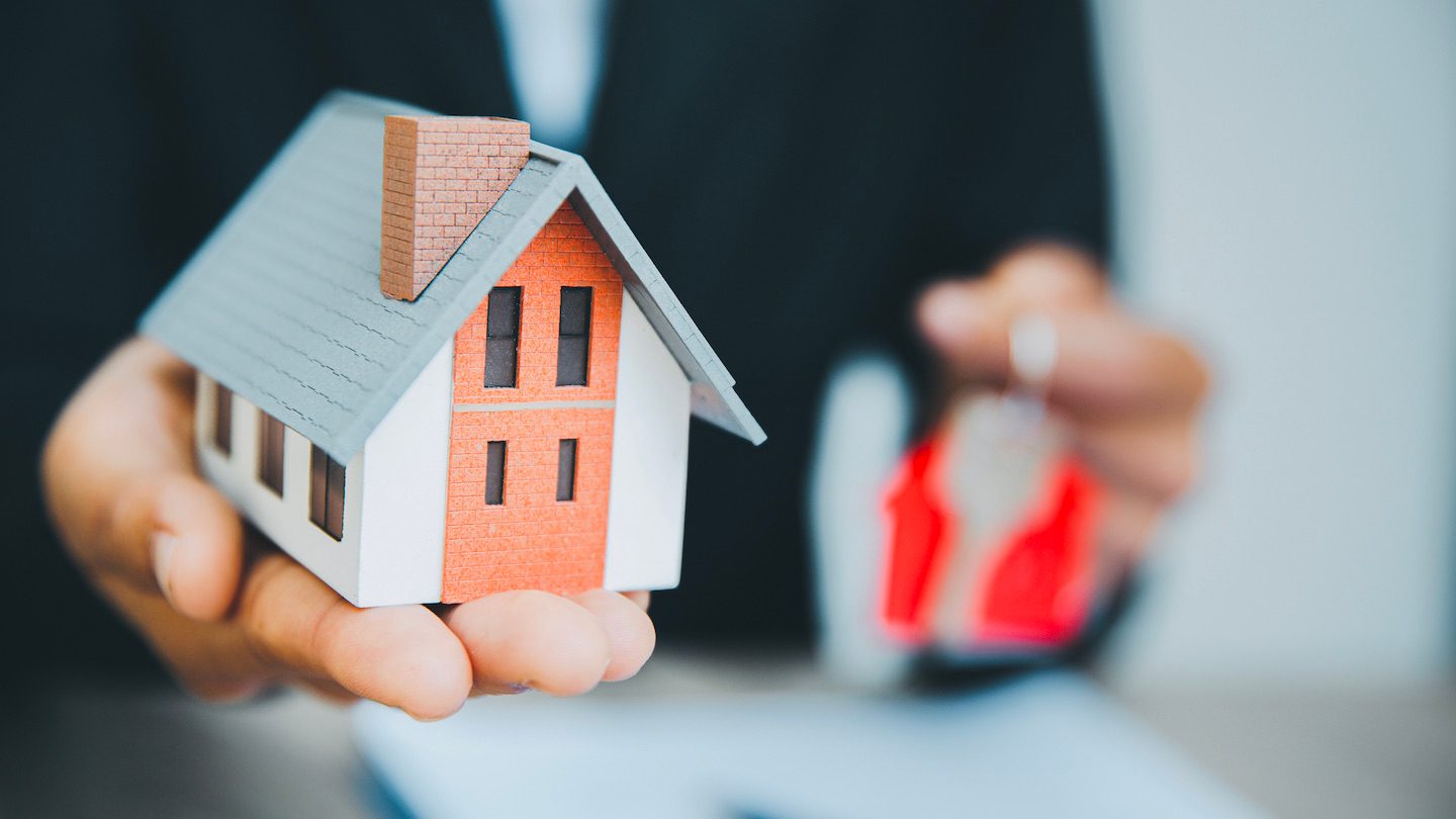 A hand holds out a model house while the other holds a key, over new first home buyer paperwork.